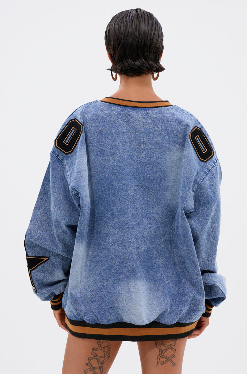 A person with short dark hair stands facing away, wearing the ALL AROUND THE WORLD DENIM SWEATSHIRT featuring black and gold patches on the shoulders and sleeves, paired with visible thigh tattoos.