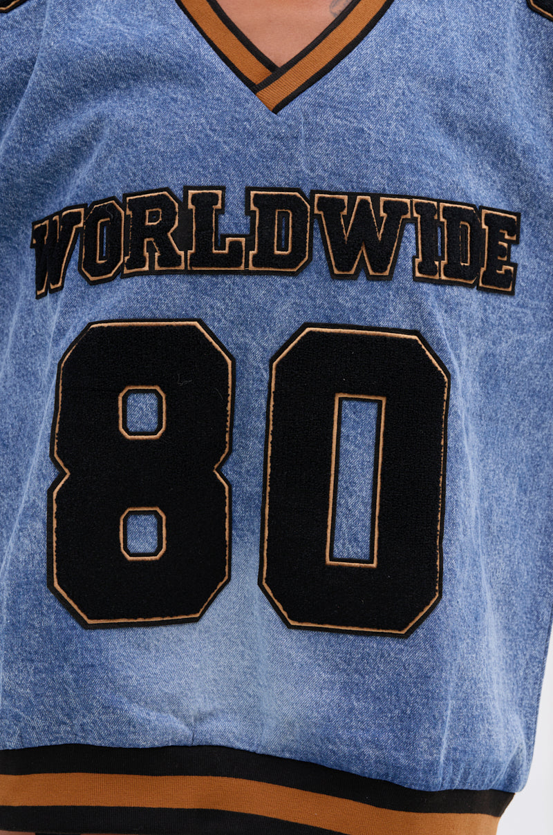 A close-up of the ALL AROUND THE WORLD DENIM SWEATSHIRT, a blue varsity-style top with "WORLDWIDE" in bold black and gold letters above a gold-trimmed 80, plus a V-neck and black-and-gold striped edging.
