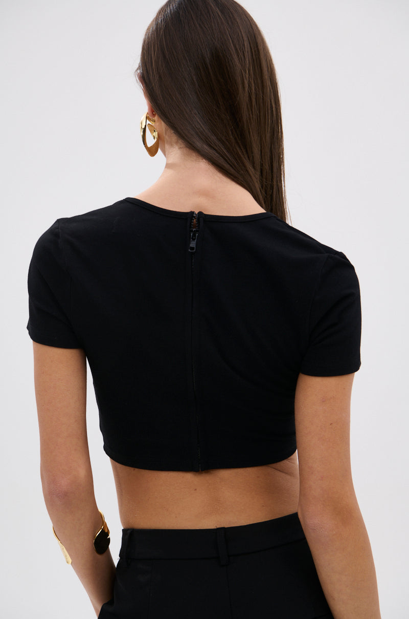 A woman with long brown hair, large hoop earrings, and a gold bracelet is shown from the back wearing the NEVER GOING TOP WITH METAL GROMMET—a black stretch crop top with a back zipper—paired with black pants. 3