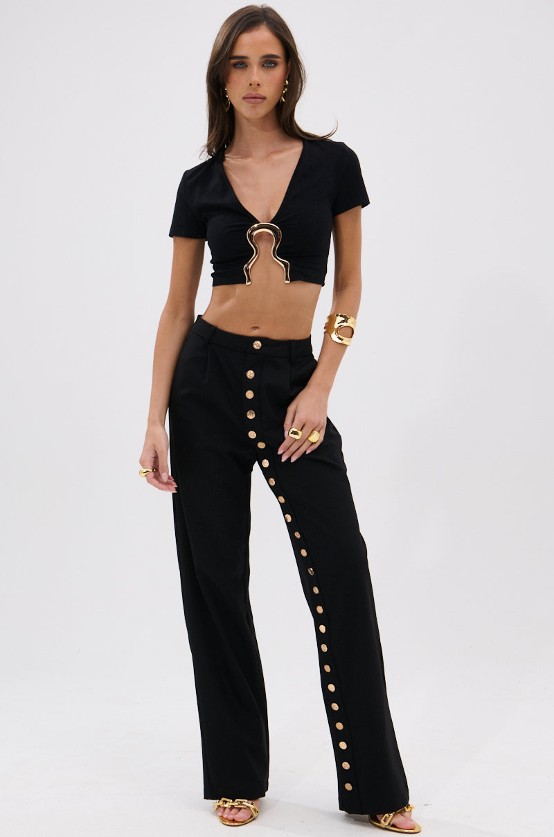 A woman stands against a plain background wearing the NEVER GOING TOP WITH METAL GROMMET, a black, fitted crop top featuring unique gold-outlined cutouts, paired with high-waisted black pants with diagonal gold buttons, accessorized with gold jewelry and sandals. 4