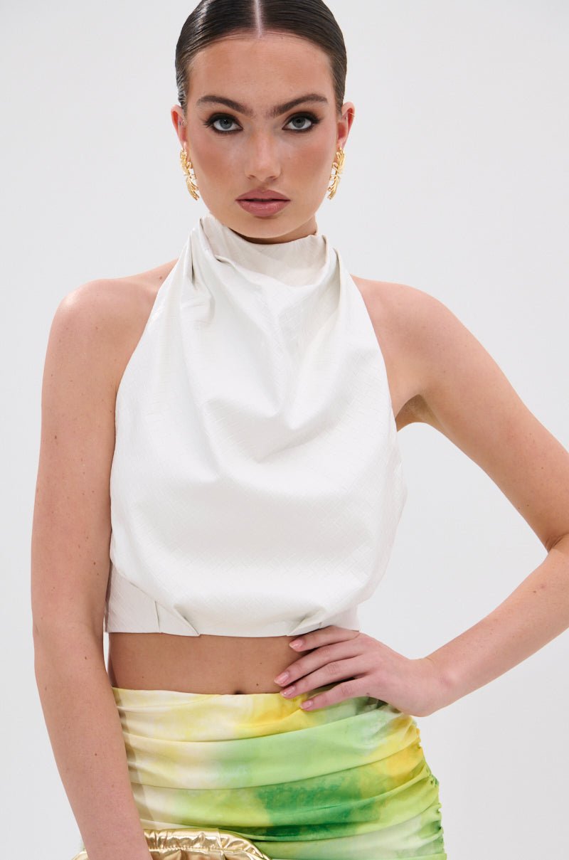 A woman with slicked-back hair wears the GOING NO WHERE HALTER BLOUSE, gold earrings, and a green and yellow tie-dye skirt, posing against a plain white background with one hand on her hip.