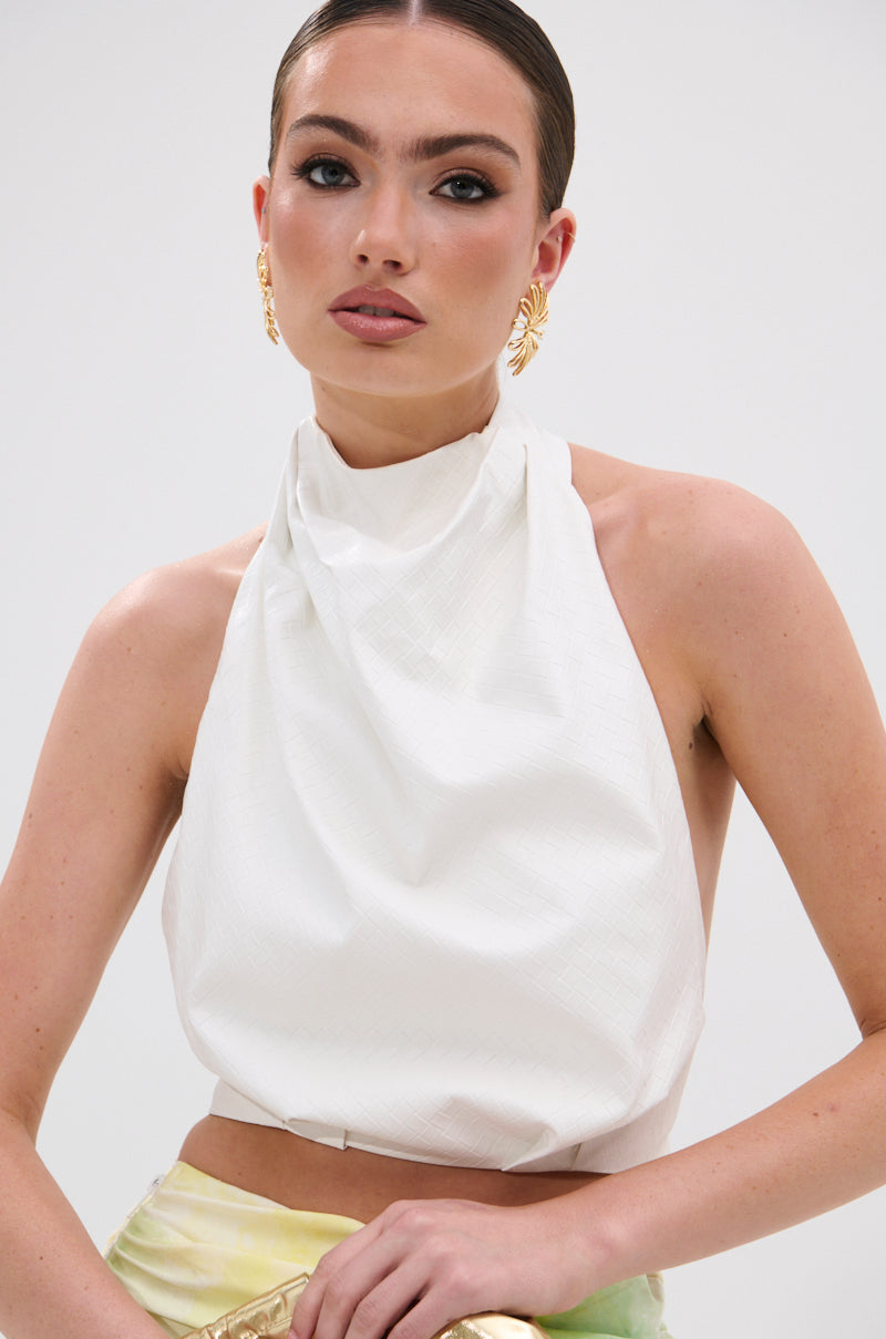 A woman with slicked-back hair wears gold statement earrings and the GOING NO WHERE HALTER BLOUSE, featuring a textured white design. She poses confidently against a plain light background, looking at the camera.
