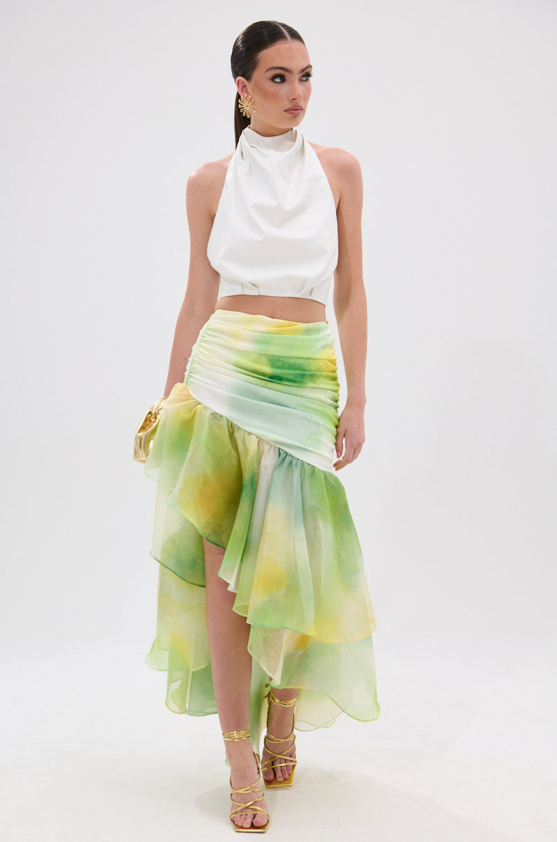 A woman wears the GOING NO WHERE HALTER BLOUSE—white, sleeveless, halter-style with an open back—paired with a green-yellow watercolor asymmetrical skirt, gold heels, and matching bag as she poses against a plain white background.