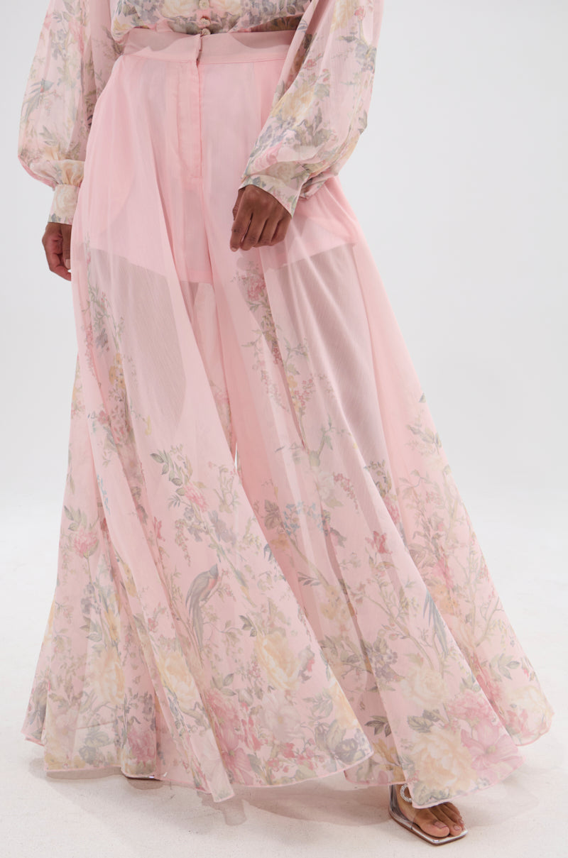 Someone wears the SIENNA FLORAL WIDE LEG TROUSER, a long, sheer, light pink trouser with soft pastel floral patterns and layered fabric underneath. Only the lower half is visible. 0