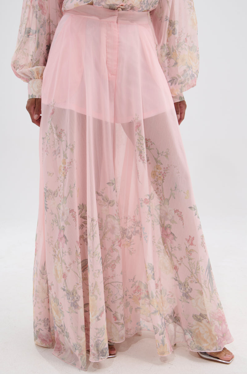 A person stands on a white background wearing the SIENNA FLORAL WIDE LEG TROUSER—sheer, light pink, floral print trousers with an inner lining—paired with a matching blouse for an airy, flowy look. 1