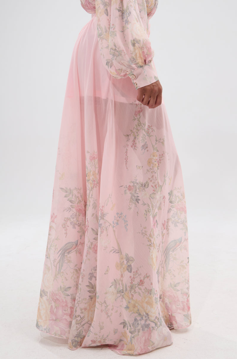 A person stands against a plain white background wearing SIENNA FLORAL WIDE LEG TROUSER, paired with a long, sheer, light pink floral dress featuring billowy sleeves and a delicate pastel flower pattern for an elegant look. 3