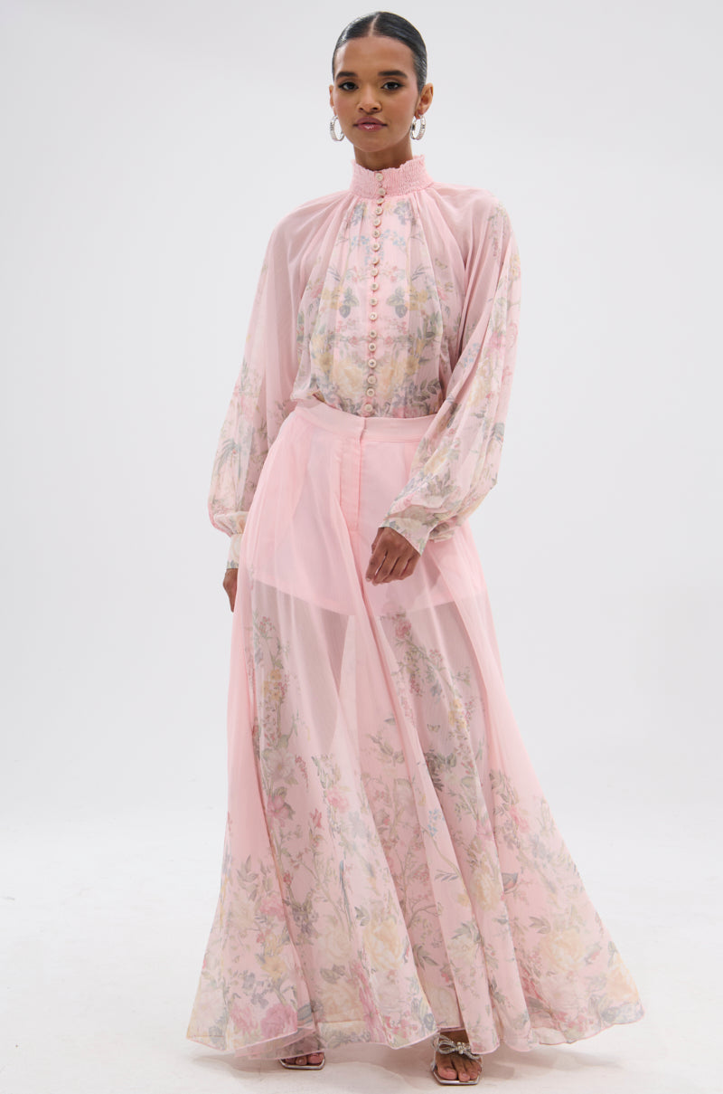 A woman styles her hair back and accessorizes with hoop earrings and heeled sandals, wearing the SIENNA FLORAL WIDE LEG TROUSER paired with a sheer, long pink top featuring floral patterns, a high ruffled collar, and puffed sleeves. 5