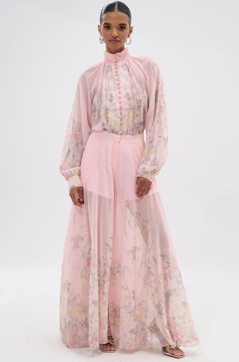 A woman wears the SIENNA FLORAL WIDE LEG TROUSER—a flowy, high-rise, chiffon pant with floral patterns—paired with a sheer light pink blouse and sandals for an effortlessly charming vintage-inspired look. 6