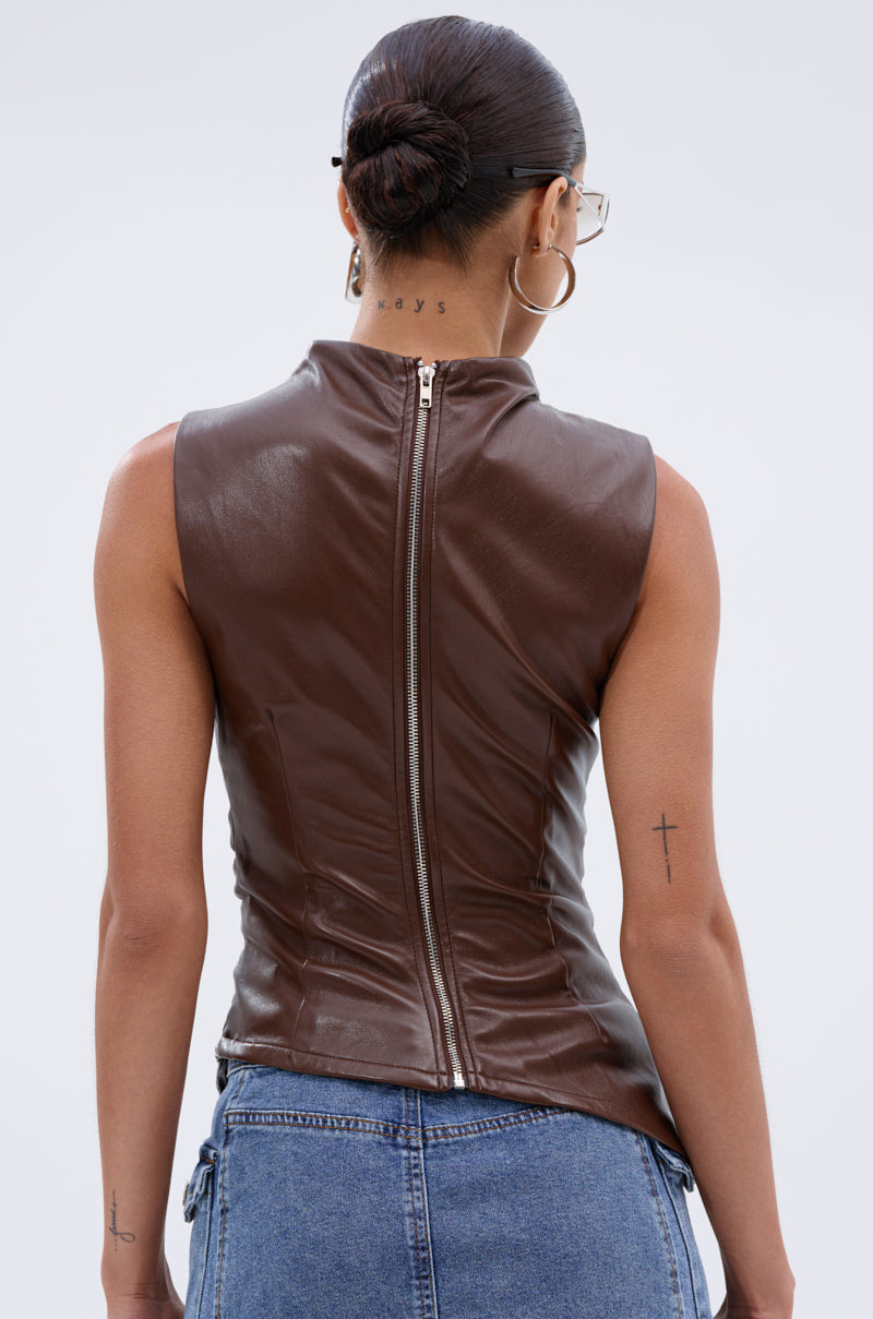A person with a neat bun, glasses, and tattoos models the LOCKED AND LOADED FAUX LEATHER BLOUSE IN CHOCOLATE BROWN, sleeveless with a center back zipper, paired with blue jeans against a plain light background. 4