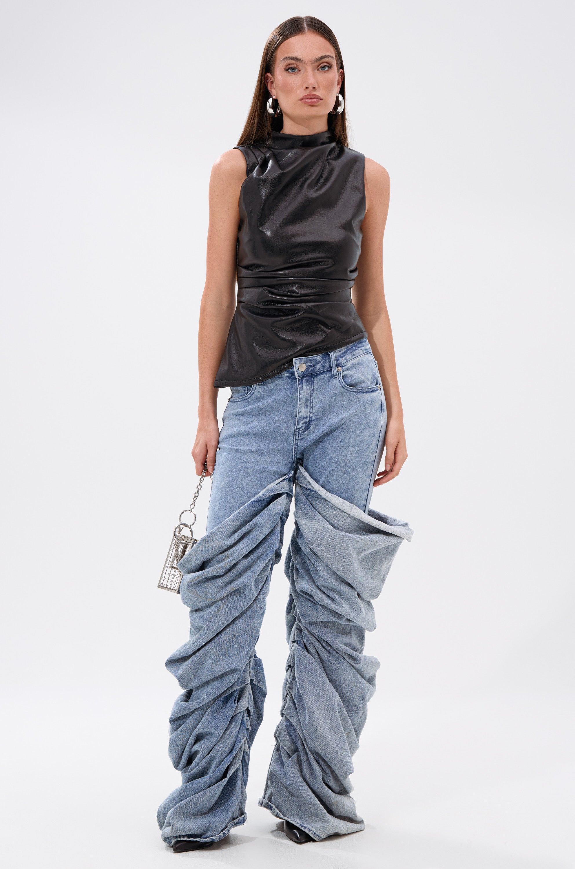 A woman models the LOCKED AND LOADED FAUX LEATHER BLOUSE with unique blue ruched wide-leg jeans, hoop earrings, and a small silver handbag, posing against a plain white background. 5