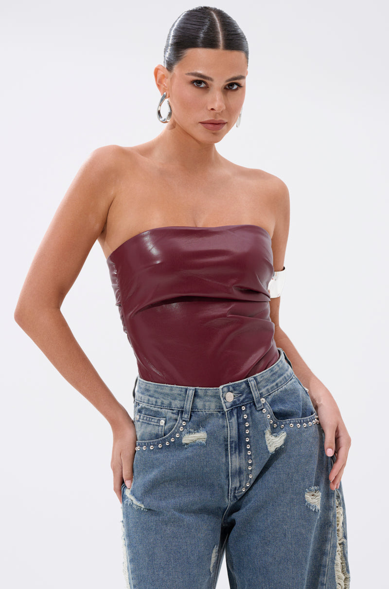 A woman poses confidently in the LUSH LIFE FAUX LEATHER PLUM BODYSUIT, styled with high-waisted light blue rhinestone jeans, slicked-back hair, and large silver hoops against a white background.
