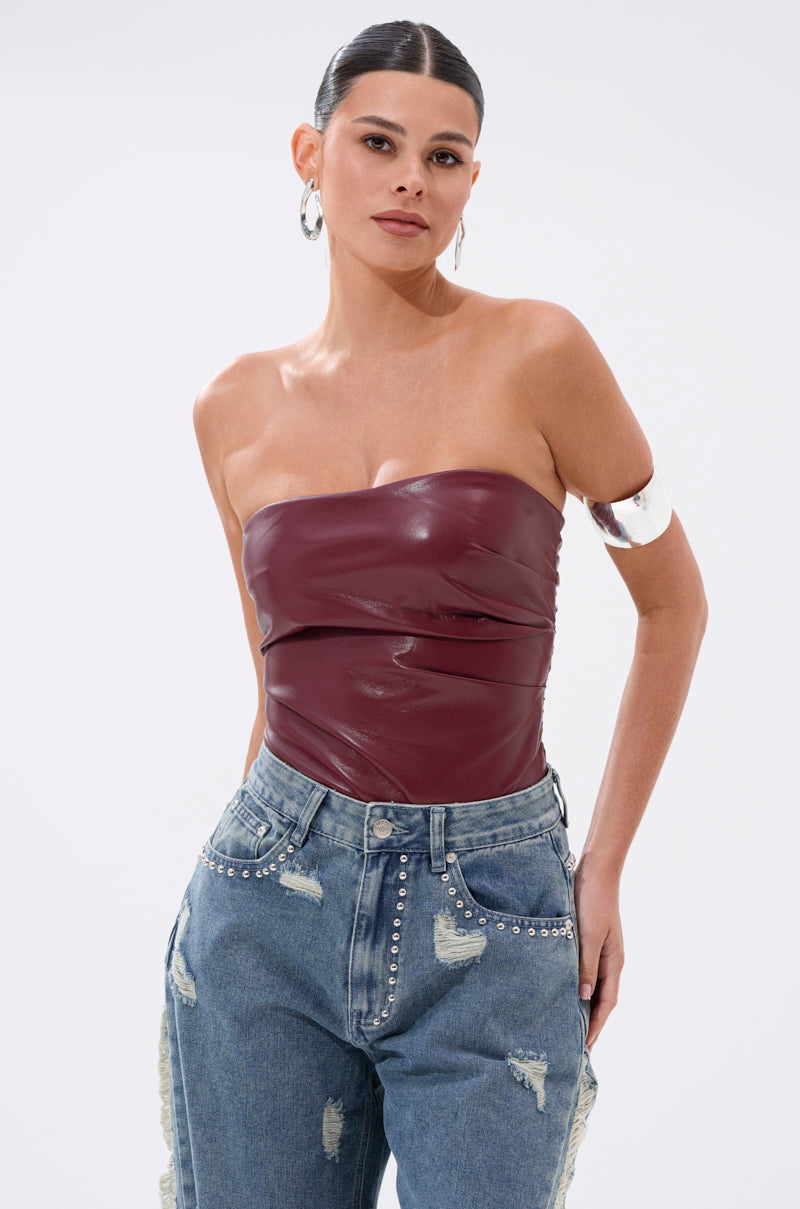 A woman with dark hair in a sleek bun wears the LUSH LIFE FAUX LEATHER PLUM BODYSUIT, paired with high-waisted distressed blue jeans featuring studded details, large hoop earrings, and a silver upper arm cuff.