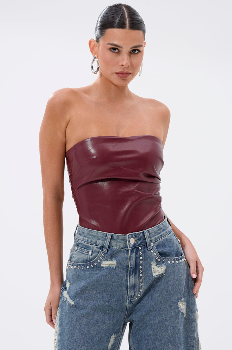 A woman wears the LUSH LIFE FAUX LEATHER PLUM BODYSUIT with high-waisted, distressed blue jeans featuring silver studs. Her hair is slicked back and she wears hoop earrings, posing confidently against a plain white background.