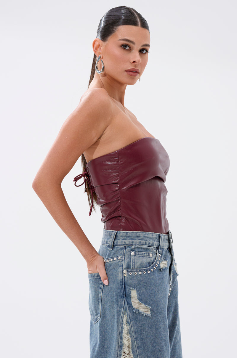 A woman with long dark hair in a ponytail models the LUSH LIFE FAUX LEATHER PLUM BODYSUIT with distressed blue studded jeans, standing sideways with one hand in her pocket against a white background.