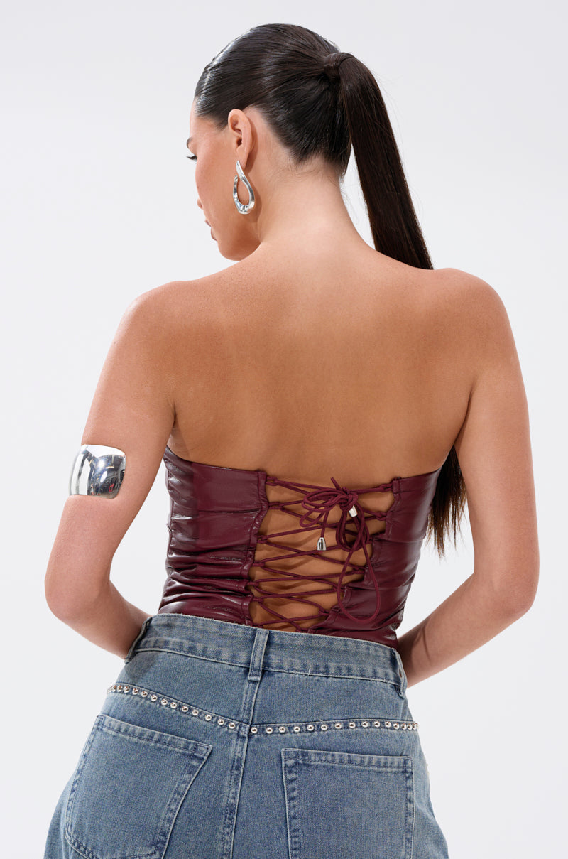 A woman with long dark hair in a ponytail is seen from behind, wearing the LUSH LIFE FAUX LEATHER PLUM BODYSUIT, high-waisted studded jeans, silver hoop earrings, and a shiny silver arm cuff.