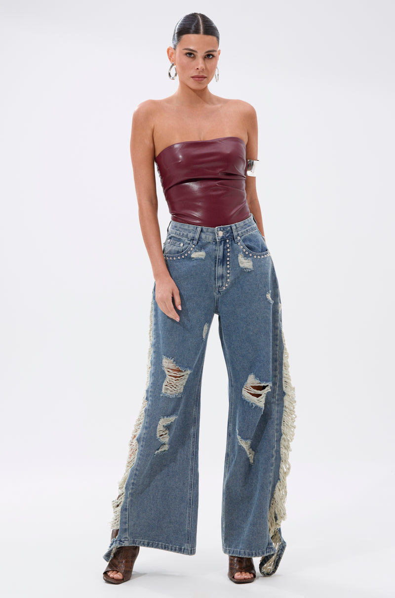 A woman stands against a white background wearing the LUSH LIFE FAUX LEATHER PLUM BODYSUIT, paired with high-waisted, wide-leg blue jeans featuring distressed rips, side fringe, stud accents, and open-toe heels.