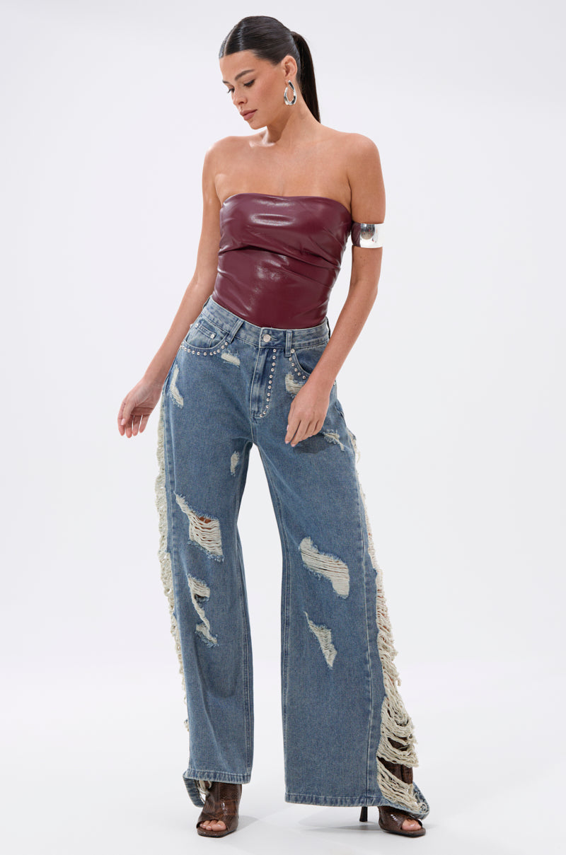A woman poses in the LUSH LIFE FAUX LEATHER PLUM BODYSUIT, paired with high-waisted, wide-leg distressed jeans with ripped sides. Her hair is pulled back and she accessorizes with hoop earrings and a metallic arm cuff.