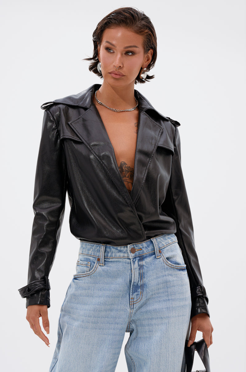 A woman with short dark hair wears the TRACIE FAUX LEATHER BLAZER BODYSUIT under a black jacket, showing part of a chest tattoo. She pairs it with light blue high-waisted jeans and holds a black bag, gazing to the side against a plain background. 1