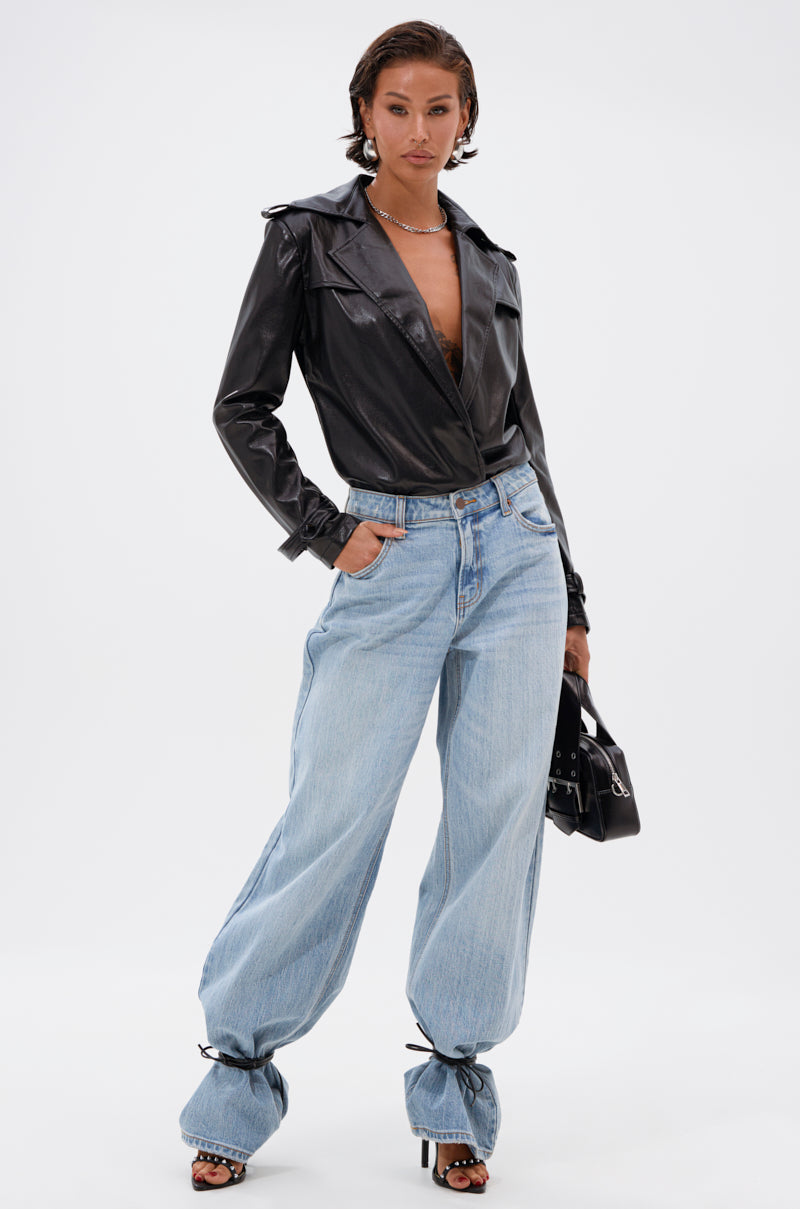 Wearing the TRACIE FAUX LEATHER BLAZER BODYSUIT, baggy light blue jeans tied at the ankles, open-toe heels, a necklace, and holding a small black handbag, a person stands against a plain background. 5