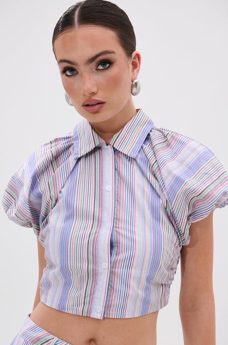 A woman with slicked-back hair wears the SUMMER TIME VIBES STRIPE BLOUSE, a colorful crop top with puffed butterfly sleeves. She pairs bold eye makeup, neutral lips, and hoop earrings against a plain white background. 1