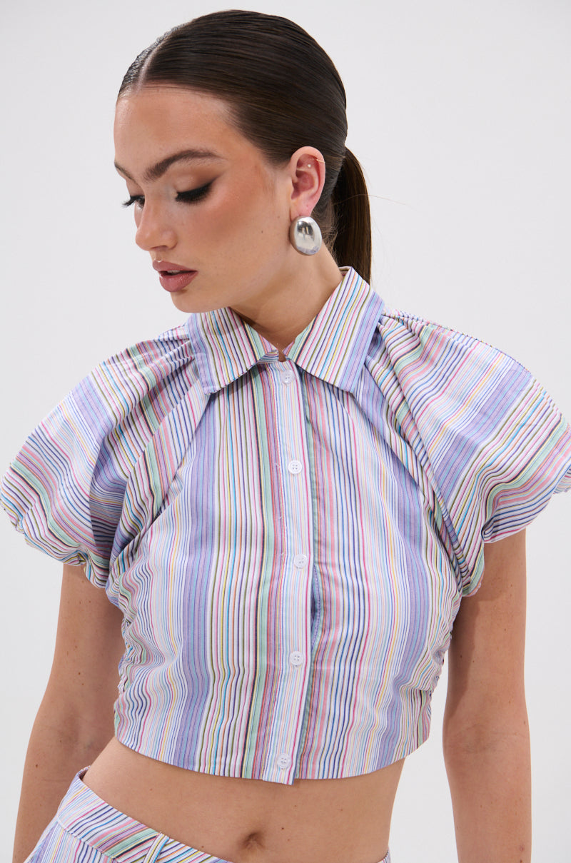 A woman with dark hair in a low ponytail wears the SUMMER TIME VIBES STRIPE BLOUSE—short-sleeved and collared with colorful vertical stripes—and large silver earrings, looking downward with her head gently turned to the side. 3