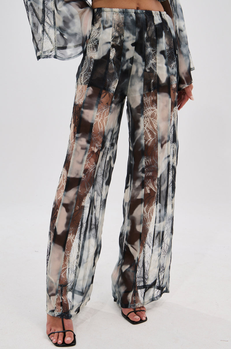 A model wears the RUBY OMBRE PALAZZO PANT, showcasing its wide-leg silhouette, tie-dye ombré print, and sheer lace panels, paired with black open-toe heels against a plain white background. 1