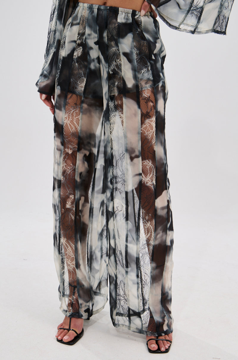 Someone wearing the RUBY OMBRE PALAZZO PANT—wide-leg, high-waisted sheer pants featuring a black, white, and gray ombré tie-dye pattern with vertical lace panels—paired with black sandals. 2