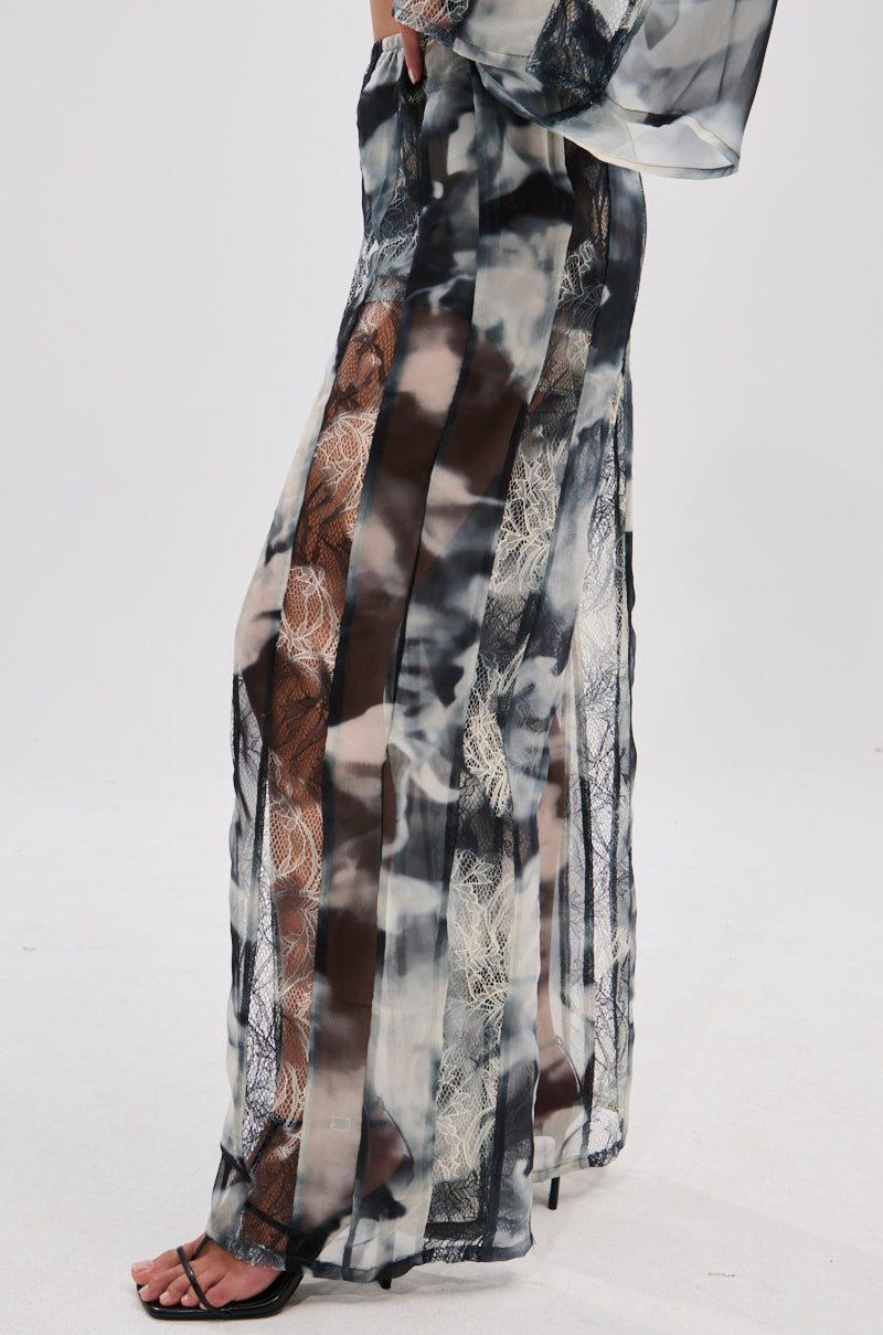A person models the RUBY OMBRE PALAZZO PANT—long, sheer, with black, white, and beige abstract patterns and vertical lace panels in a wide-leg silhouette—paired with black heeled sandals against a plain white background. 3