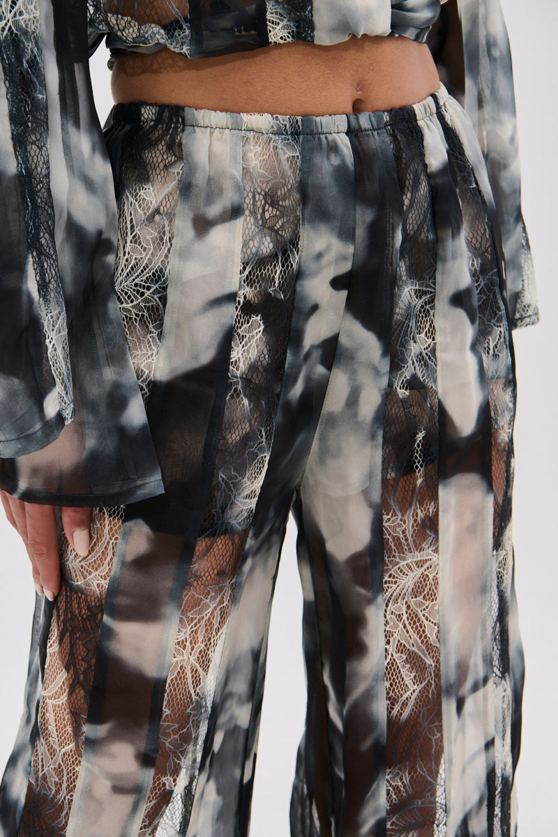 A close-up of a person wearing the RUBY OMBRE PALAZZO PANT, featuring sheer, wide-leg ombré pants with black, gray, and white tie-dye stripes and lace details. The matching long-sleeve top is partially visible. 7