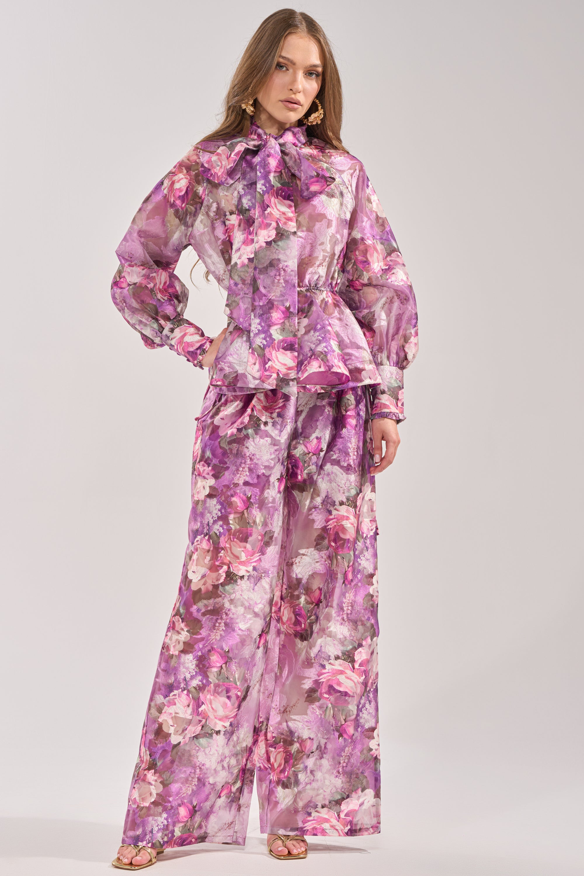 A woman models the KEEP IT FRESH PALAZZO PANT, a sheer, purple floral wide-leg style. She pairs it with a peplum top featuring long sleeves and a bow neck, accessorized with gold earrings and sandals for a resort-ready look. 0