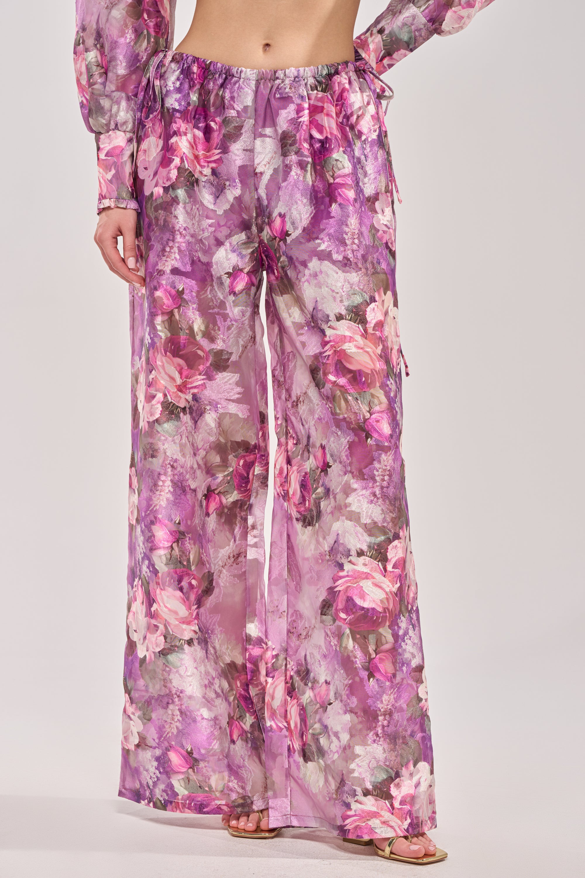 Someone stands against a plain light background wearing KEEP IT FRESH PALAZZO PANT—purple sheer floral pants with a loose, wide-leg fit and drawstring waist, perfect for resort wear. 1