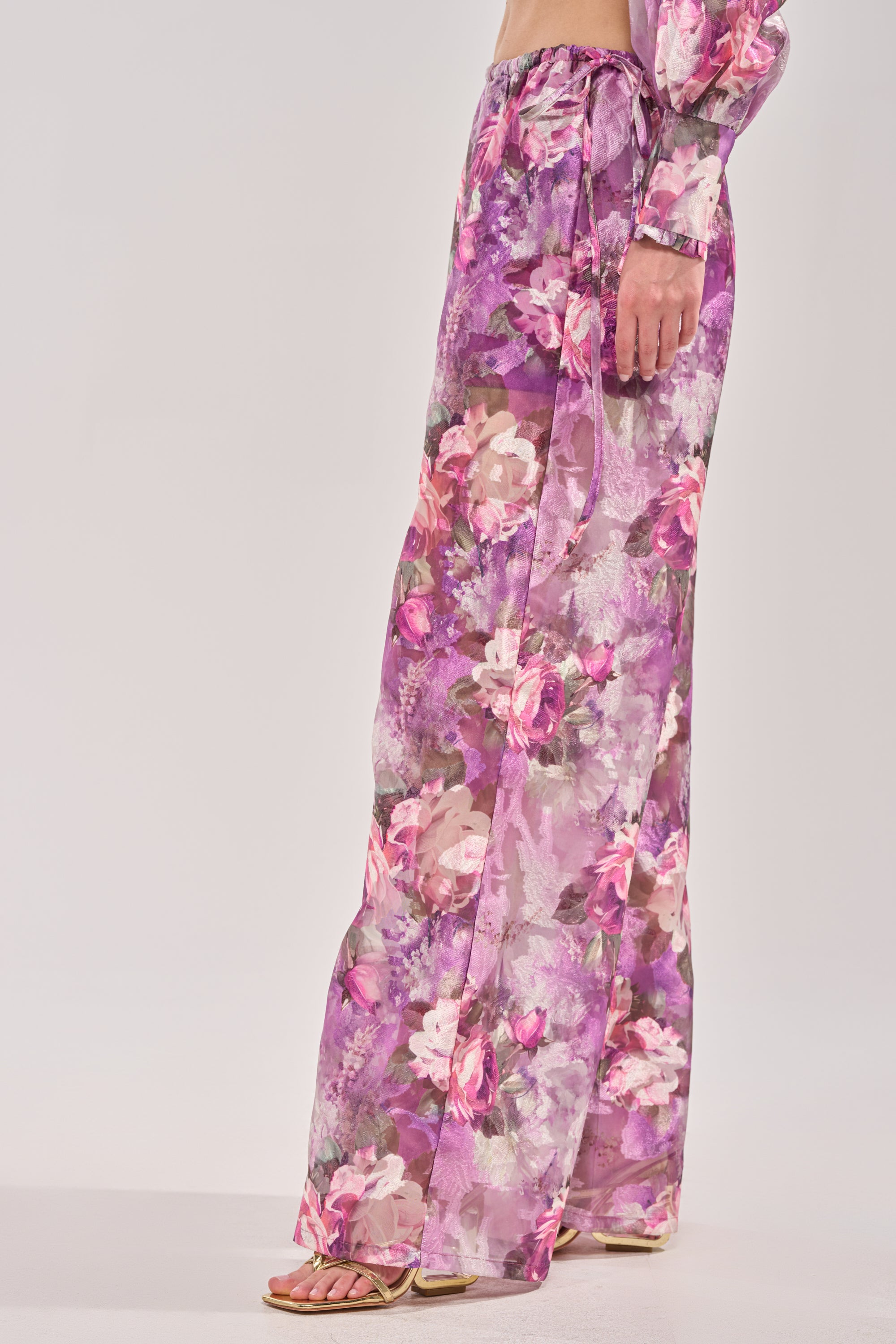 Someone stands against a light background wearing the KEEP IT FRESH PALAZZO PANT, featuring a purple floral print and high-rise elastic waistband, paired with a matching crop top and sandals. 2