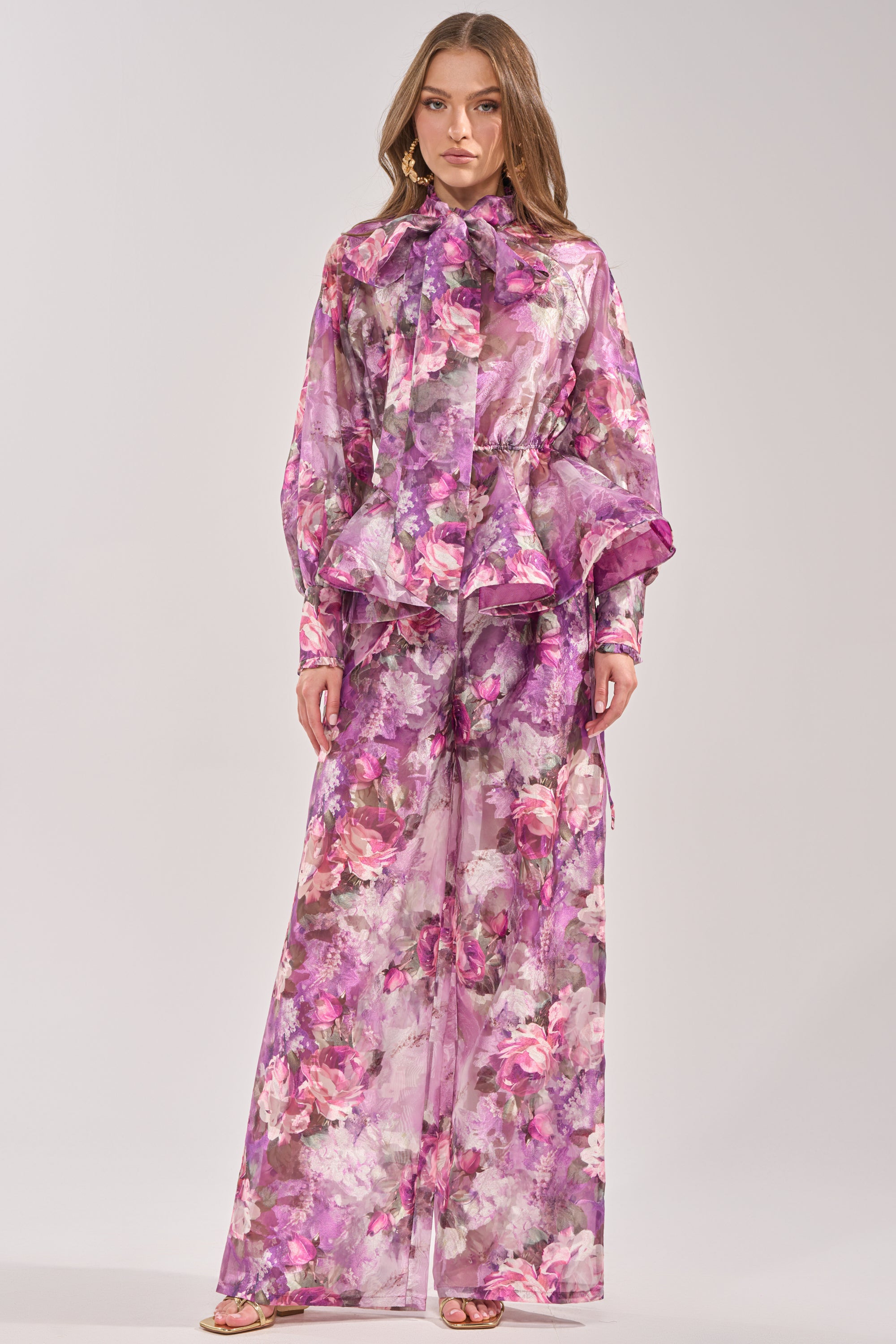 A woman poses against a plain background in the KEEP IT FRESH PALAZZO PANT, featuring a pink, purple, and white sheer floral pattern—perfect resort wear—styled with a matching long-sleeve blouse, loose hair, and neutral makeup. 4
