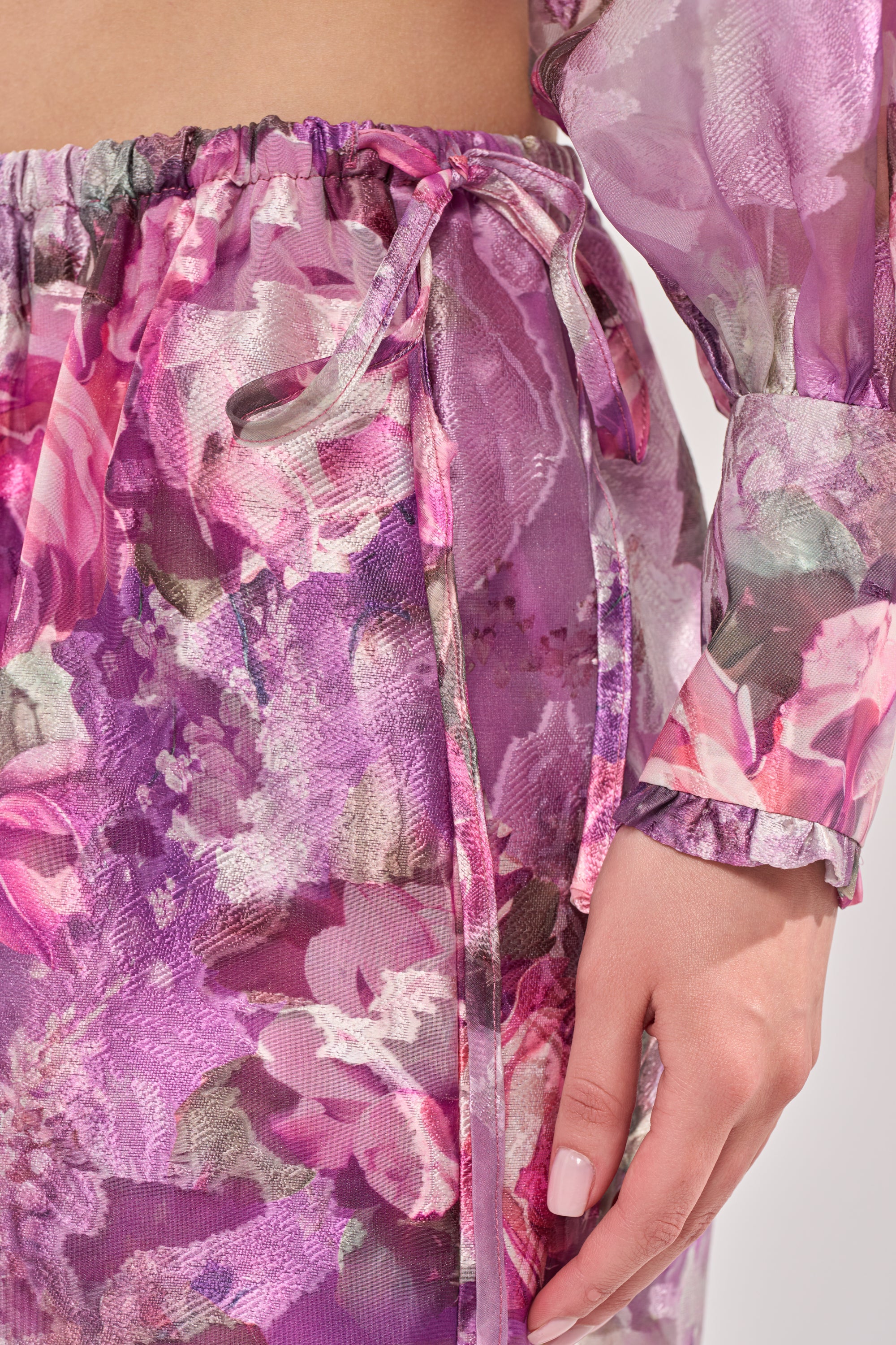 A close-up of a person wearing the KEEP IT FRESH PALAZZO PANT, styled as resort wear with a drawstring waist and matching top. The sheer purple floral pants are showcased, with the person's hand resting at their side. 5