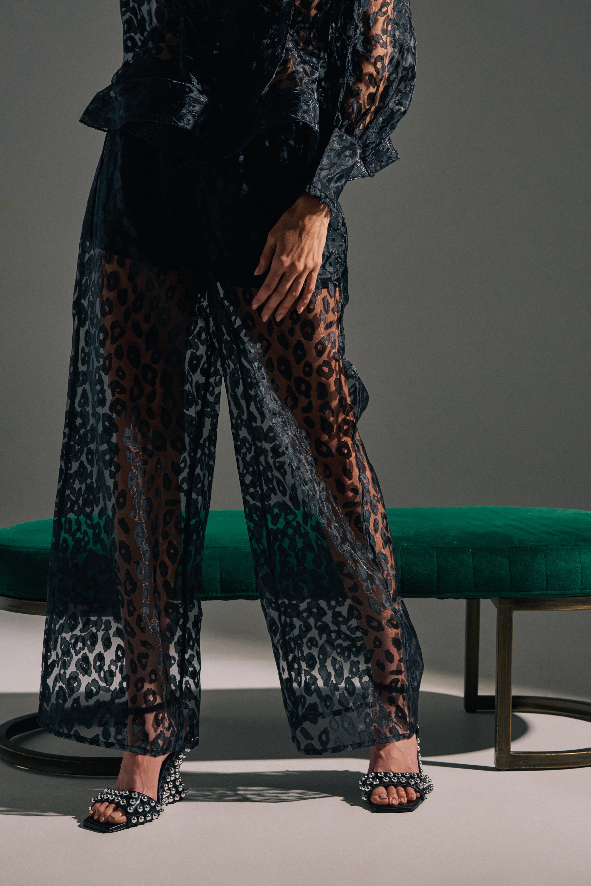 Wearing CHEETAH SISTA PALAZZO PANT and a matching top, a person stands beside a green velvet bench in studded open-toed shoes, set against a neutral, minimal background.