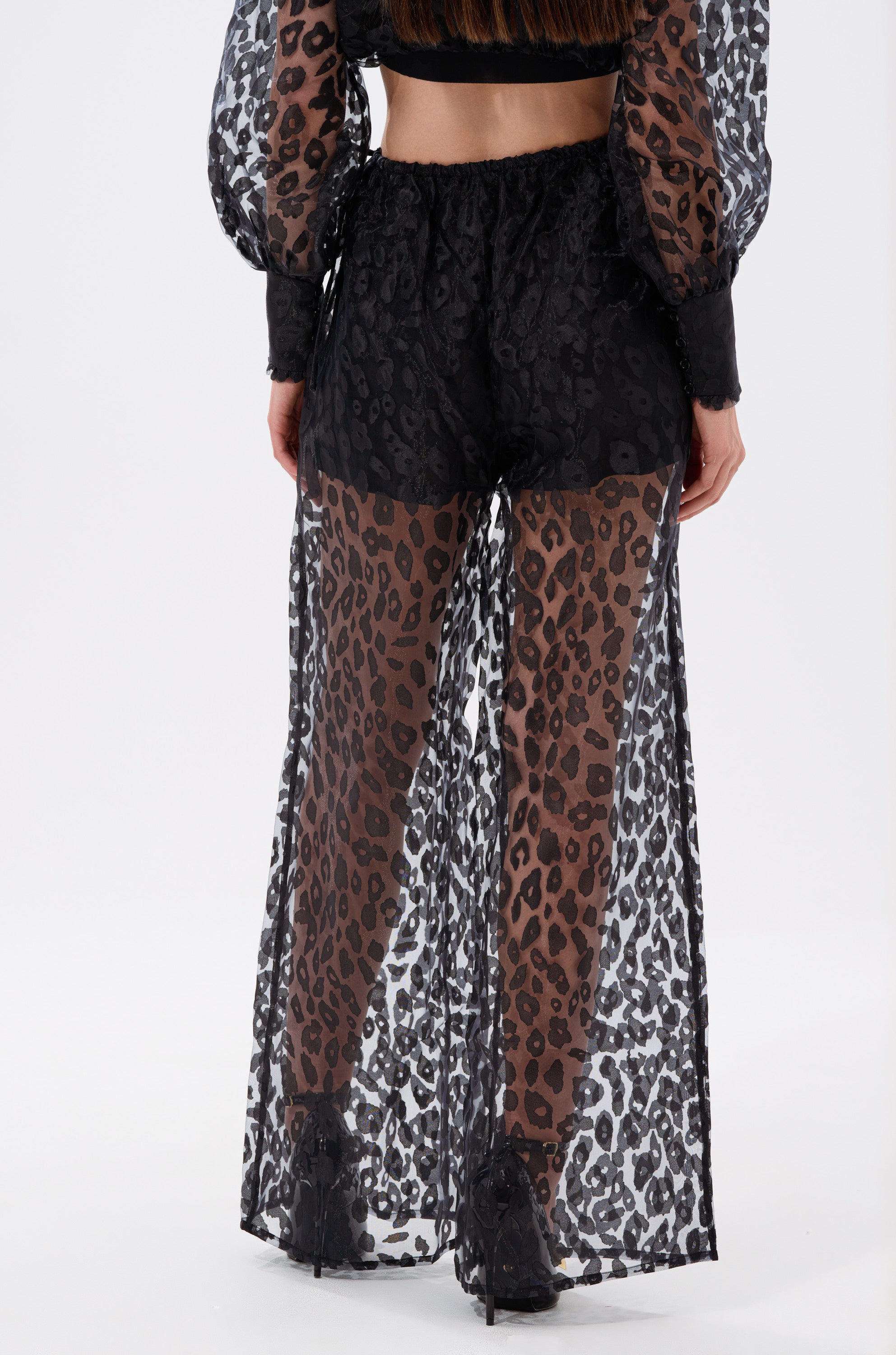 A person shown from the back wears the CHEETAH SISTA PALAZZO PANT—sheer chiffon, high-rise, wide-leg pants with a leopard print pattern—over brown leopard shorts and black high-heeled shoes.