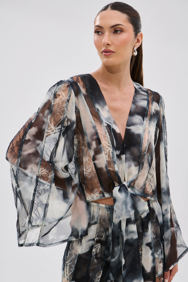 A woman with long brown hair in a low ponytail wears the RUBY OMBRE TIE FRONT BLOUSE—sheer chiffon with black, white, and beige abstract patterns, lace panels, wide sleeves, and a front knot—paired with matching pants and pearl drop earrings.
