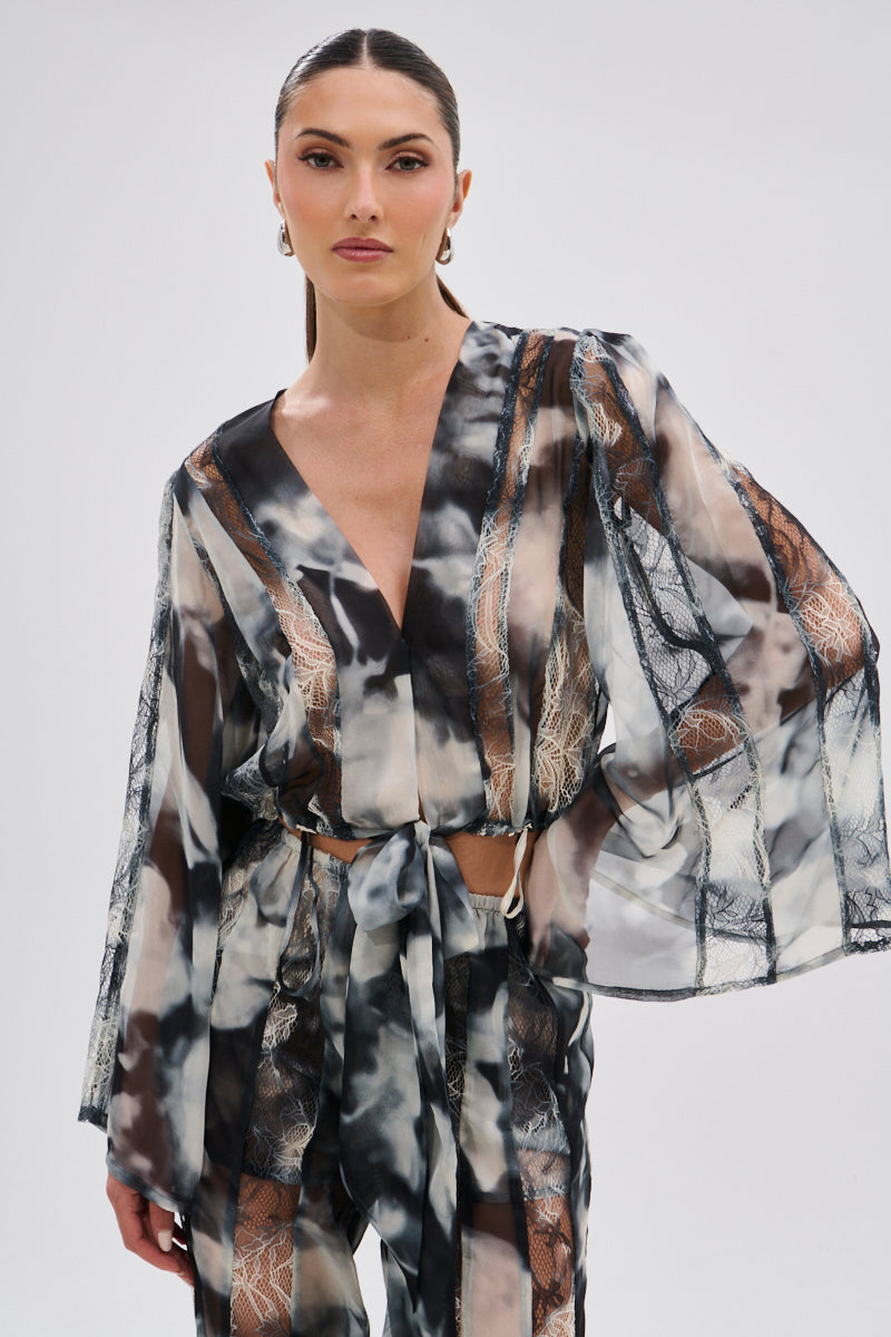 A woman with slicked-back hair models the RUBY OMBRE TIE FRONT BLOUSE, showcasing sheer chiffon fabric, wide sleeves, lace-like black and white details, and a deep V-neck as she poses against a light background.