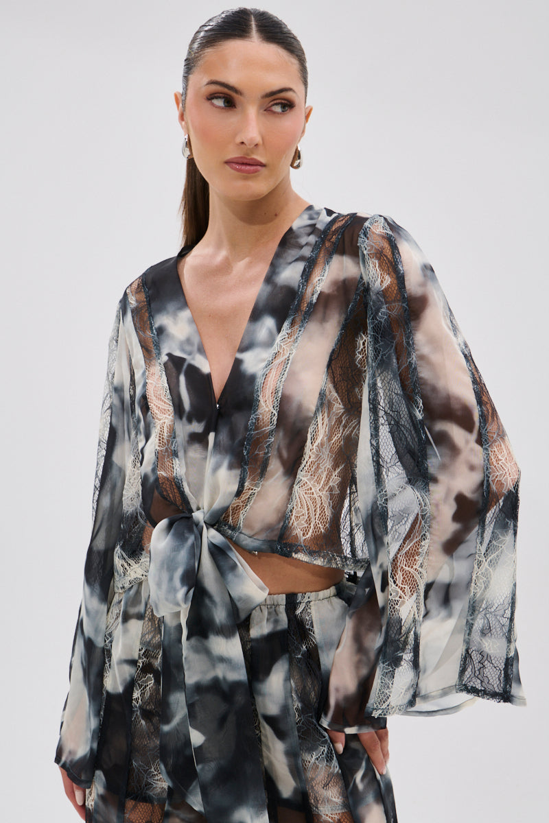 A woman with slicked-back hair wears the RUBY OMBRE TIE FRONT BLOUSE, a sheer chiffon, long-sleeved top with an abstract black, white, and beige pattern. She gazes sideways with a neutral expression against a plain background.