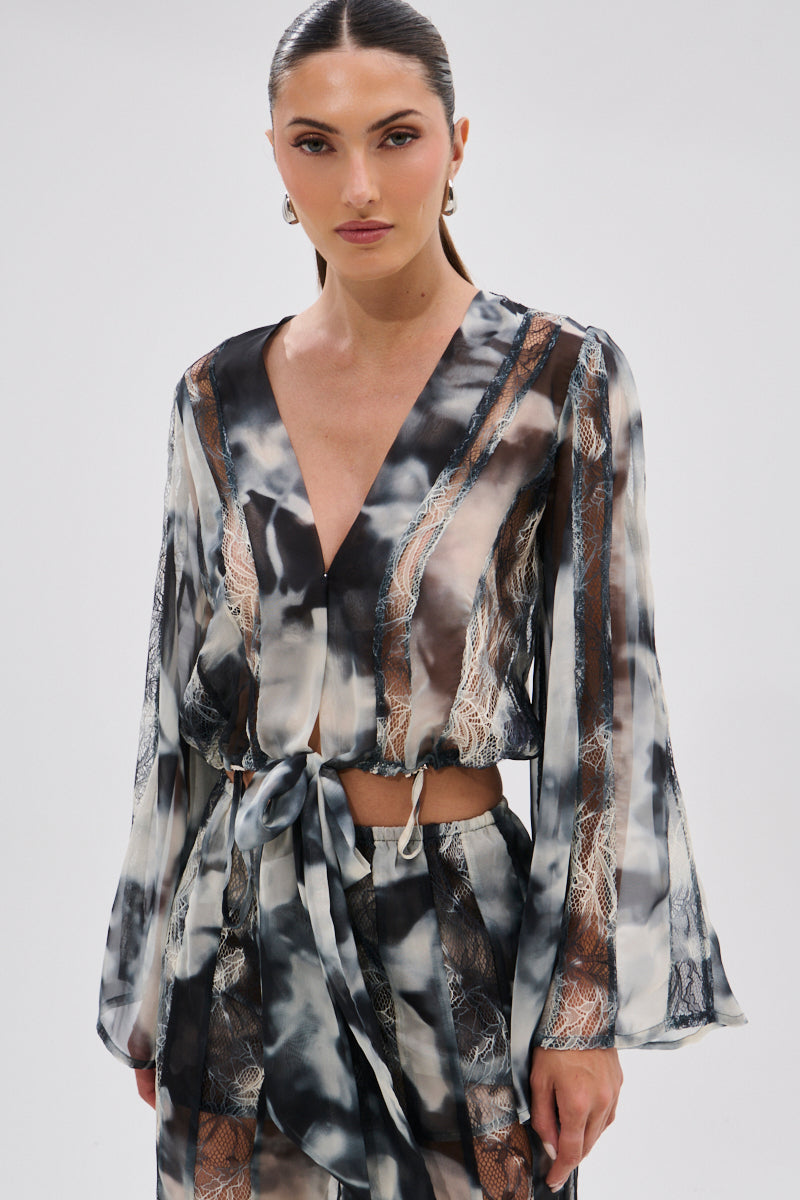 A woman with dark hair tied back wears the RUBY OMBRE TIE FRONT BLOUSE—a long-sleeve chiffon top with a V-neck, tie-front, lace panels, and a black, white, and beige abstract pattern—standing against a plain light background.