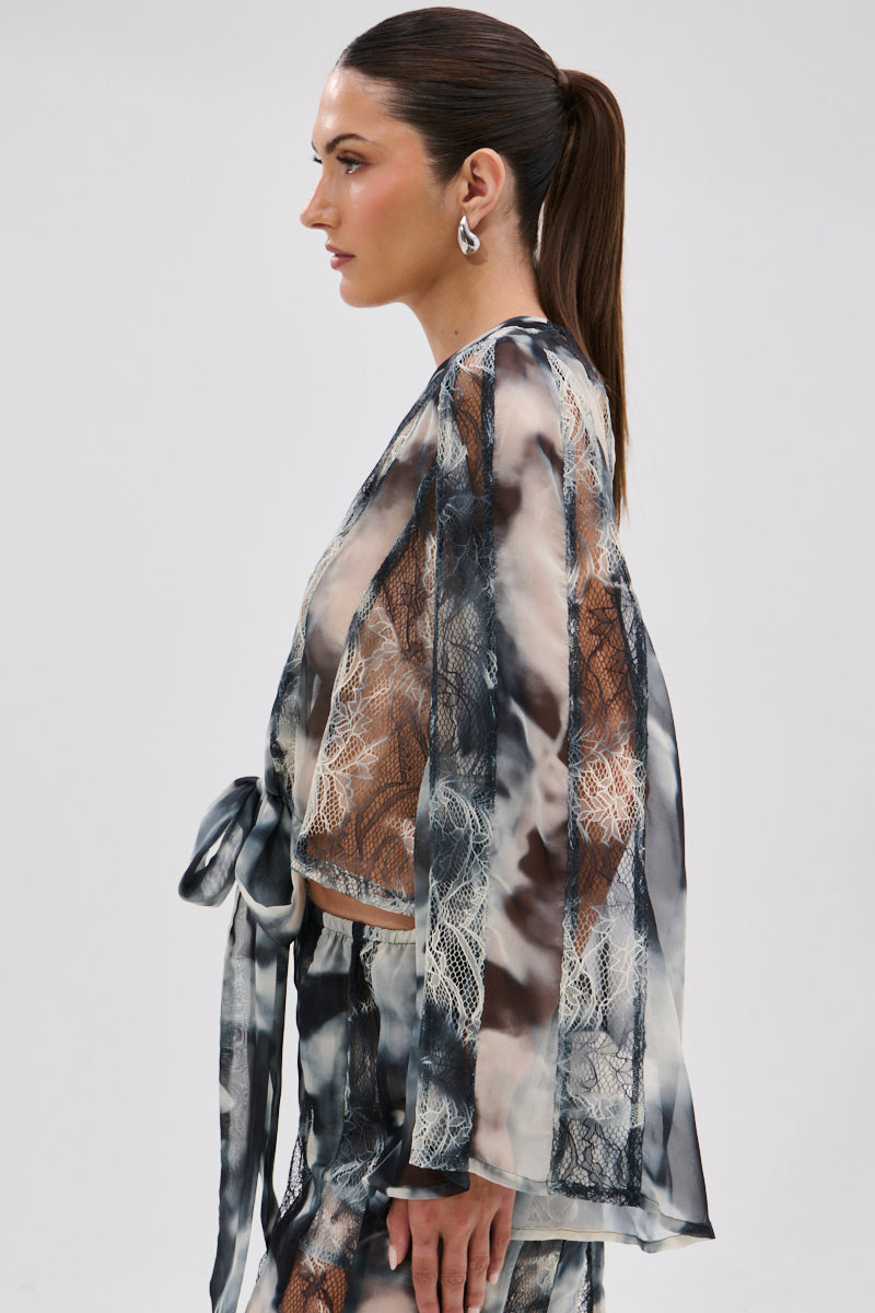 A woman with long brown hair in a ponytail stands in profile, wearing the RUBY OMBRE TIE FRONT BLOUSE—a sheer chiffon kimono-style top with lace panels and a belt—paired with matching pants featuring black, white, and gray abstract designs.