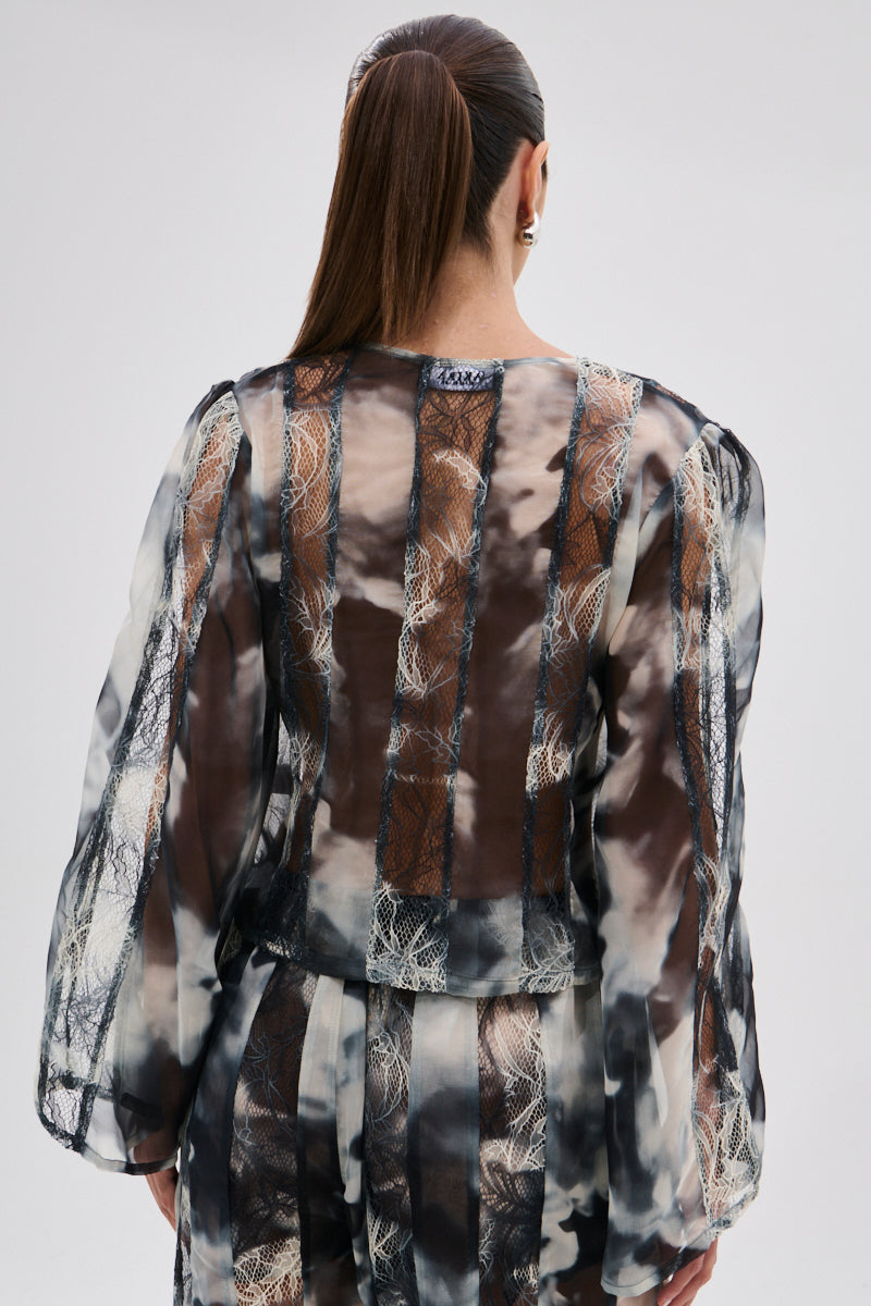 A woman with a sleek ponytail, seen from behind, wears the RUBY OMBRE TIE FRONT BLOUSE—a sheer chiffon top featuring long sleeves and ombre-patterned panels in black, white, and brown. She stands against a plain light background.