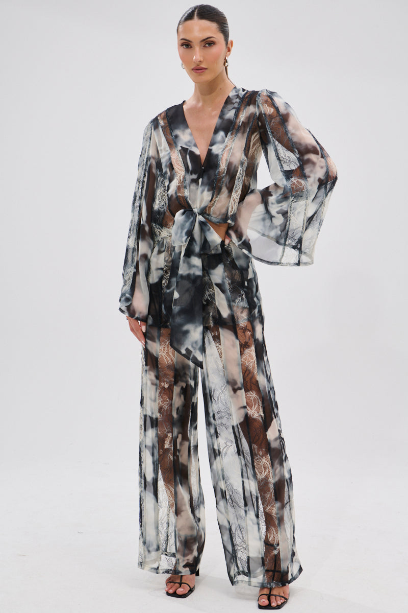 A woman stands against a white background wearing the RUBY OMBRE TIE FRONT BLOUSE—a sheer chiffon, long-sleeve, tie-front top—paired with matching wide-leg pants in a black, white, and gray abstract pattern and black strappy heels.