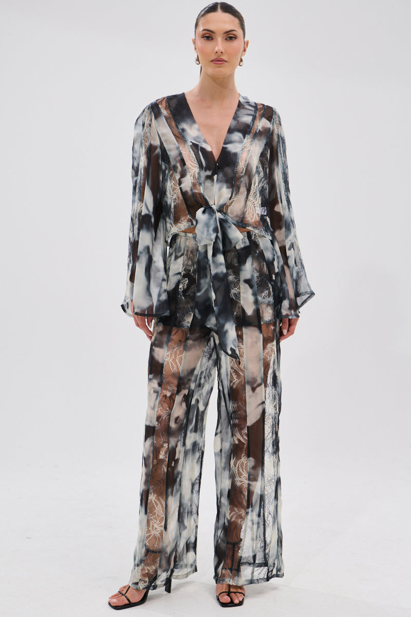 A woman poses against a white background wearing the RUBY OMBRE TIE FRONT BLOUSE, a sheer black and white chiffon top with lace panels, deep V-neckline, long sleeves, and waist tie, styled with wide-leg pants and black strappy heels.