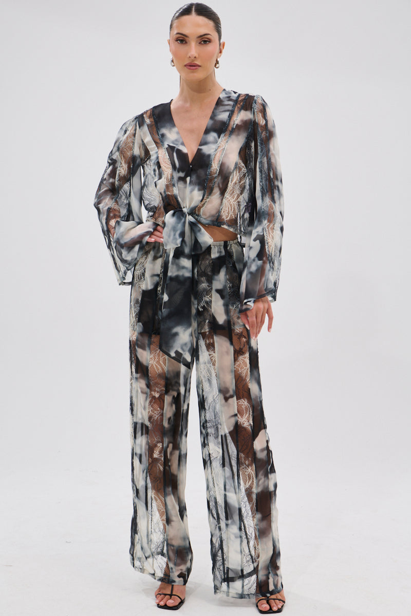 A woman with slicked-back hair wears the RUBY OMBRE TIE FRONT BLOUSE featuring lace panels, paired with matching wide-leg pants in an abstract black, white, and brown pattern and black heels against a white background.