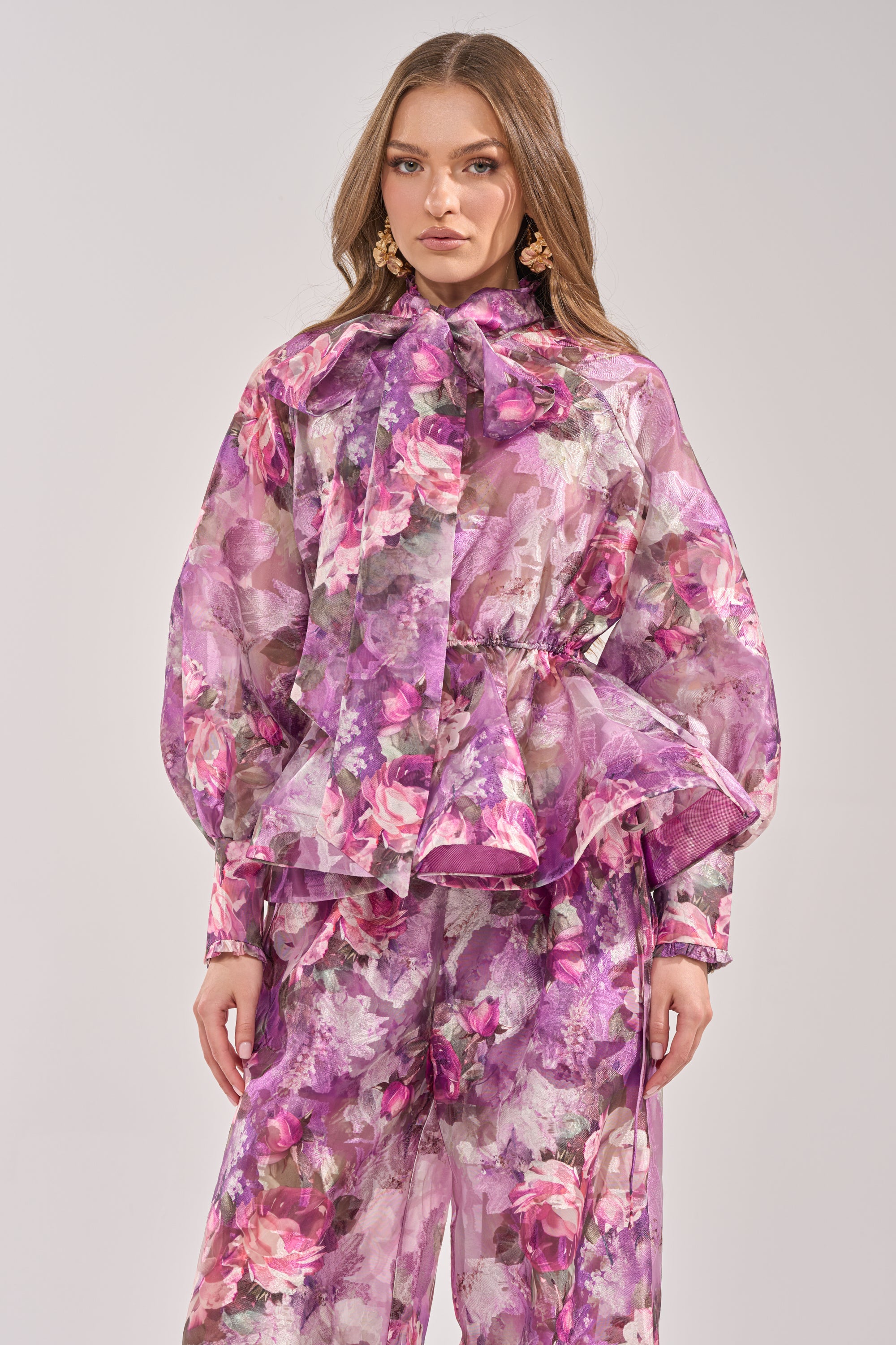 A woman wears the KEEP IT FRESH FLORAL BLOUSE, a sheer purple top with floral print, long sleeves, and a statement neck bow, paired with matching pink and purple wide-leg pants against a plain light background. 0