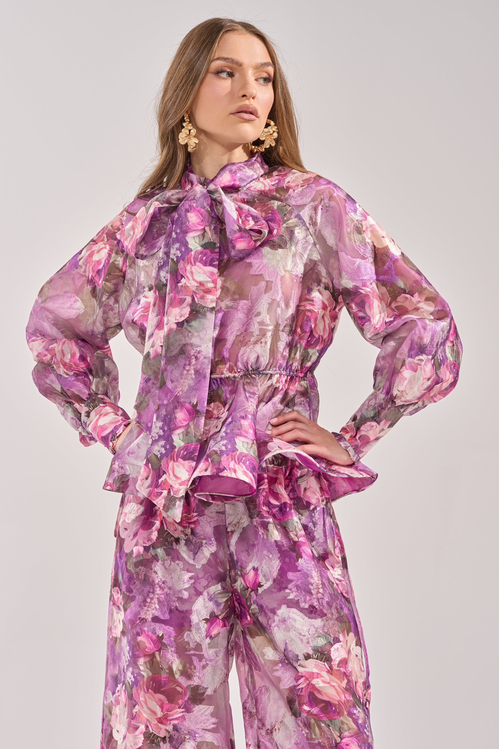 A woman poses in the KEEP IT FRESH FLORAL BLOUSE, a long-sleeve top with a ruffled mock neck, large bow, and ruffled hem in pink, purple, and green. She pairs it with matching wide-leg pants, statement earrings, and wavy hair. 1