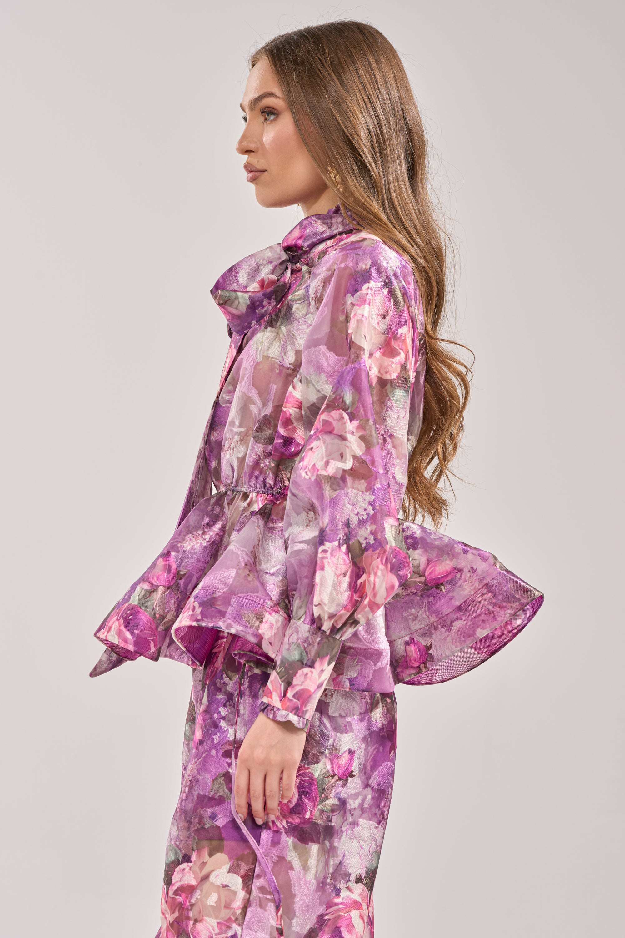 Woman in profile wears the KEEP IT FRESH FLORAL BLOUSE—sheer purple with floral print, ruffled peplum waist, and mock neck—paired with a matching skirt. She has long, wavy brown hair and stands before a plain light background. 2