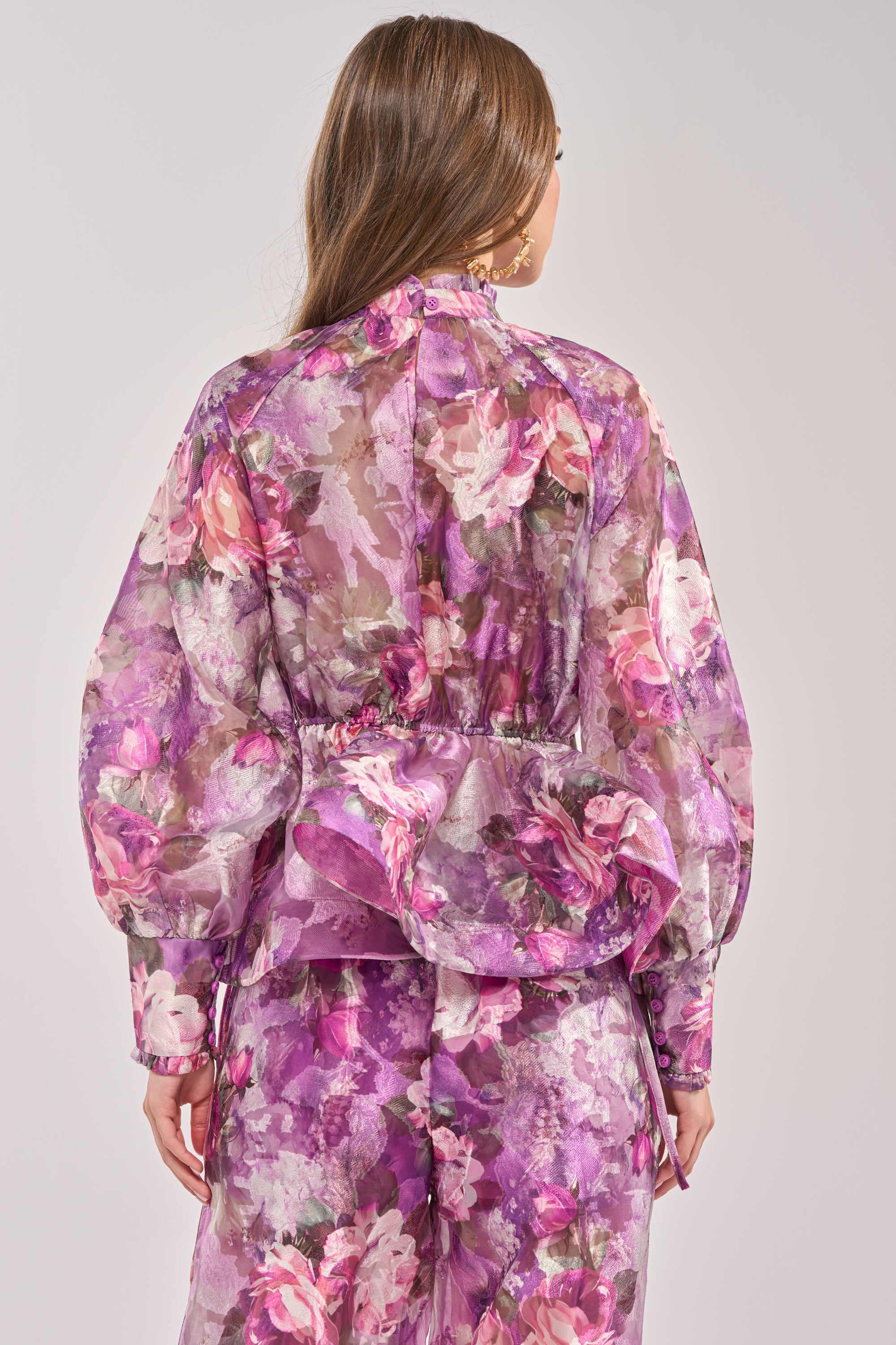 A woman with long brown hair is seen from behind wearing the KEEP IT FRESH FLORAL BLOUSE, a sheer purple top with a ruffled mock neck and peplum hem, paired with matching floral print pants in purple, pink, and white. 3