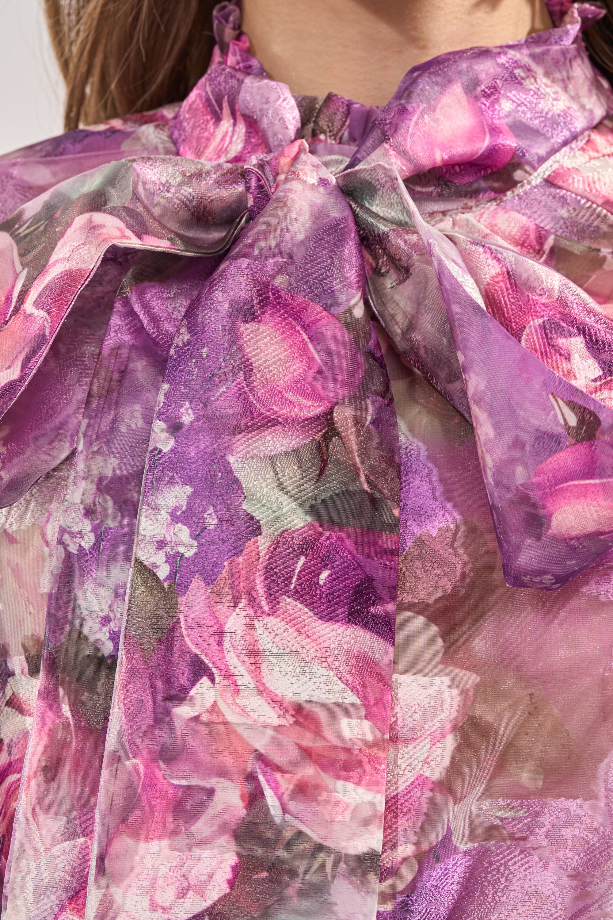 Close-up of the KEEP IT FRESH FLORAL BLOUSE, a sheer purple top with pink, purple, and green floral print. Features a tied bow at the ruffled mock neck and elegant ruffle details for a refined look. 6